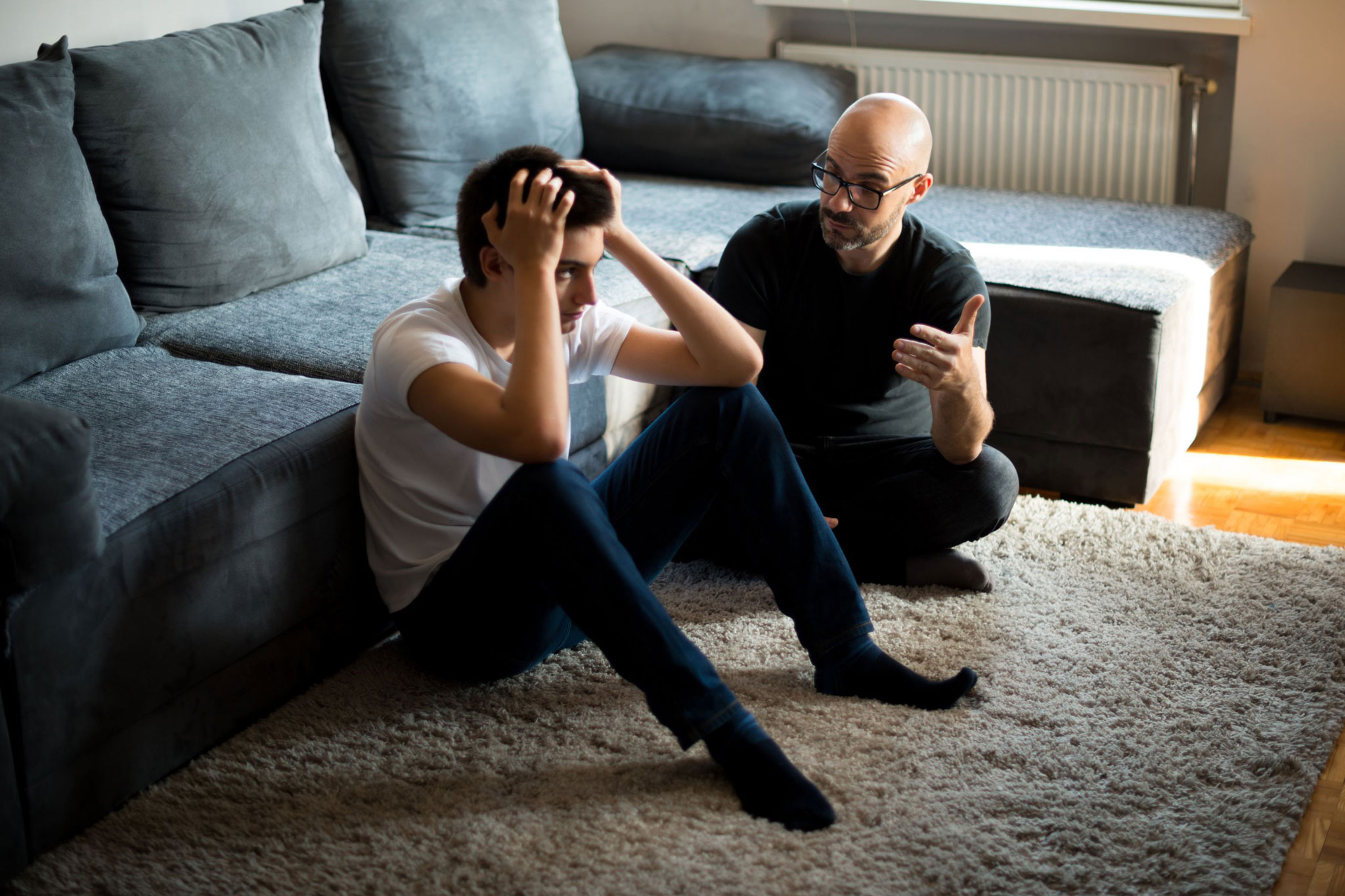 A father sits on the floor with his teenage son during a serious talk at home; the son looks distressed while the parent gestures supportively, illustrating a difficult but necessary conversation about sextortion prevention